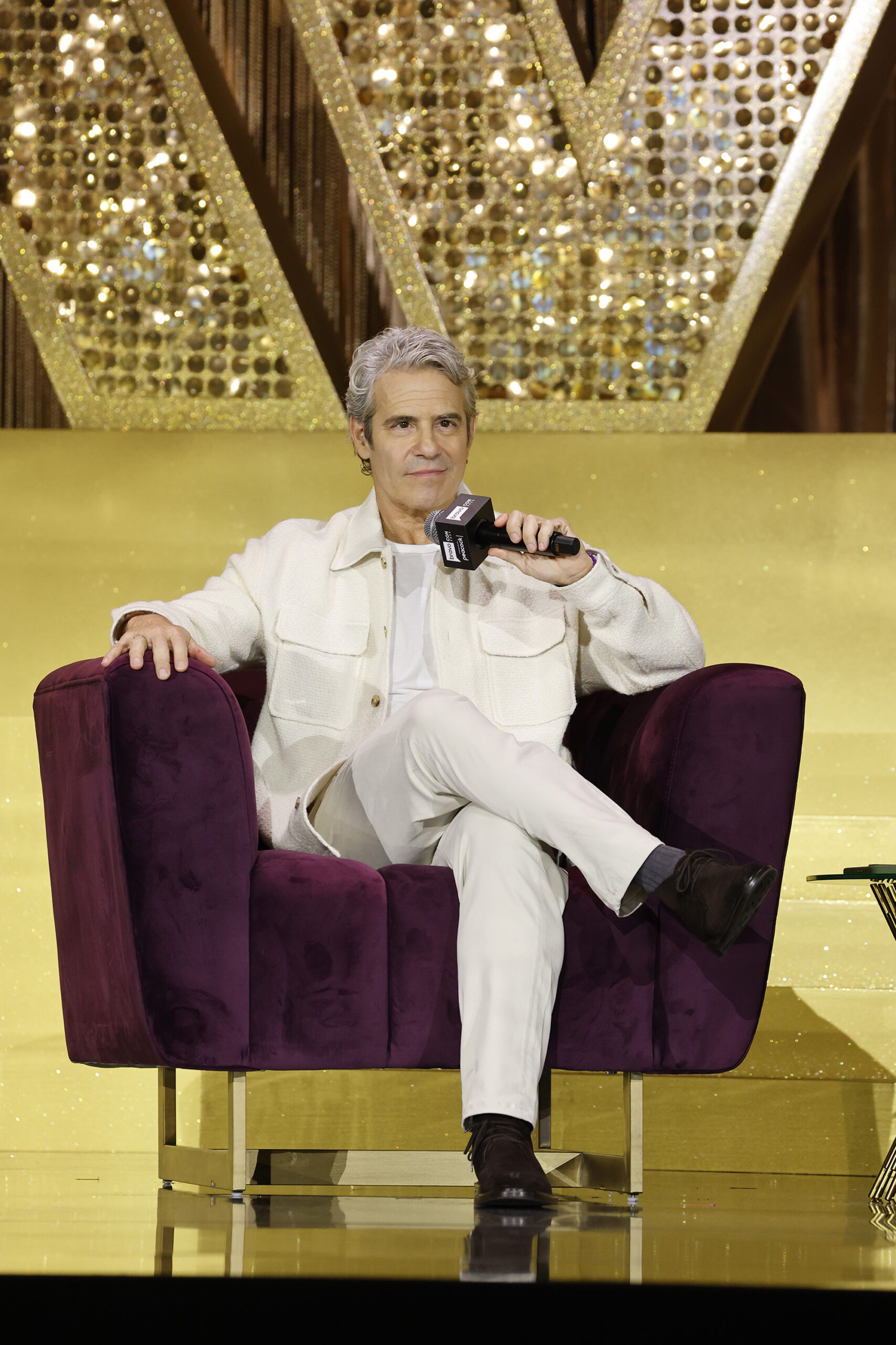 Andy Cohen sitting in a purple armchair holding a microphone on the BravoCon Gold Stage.