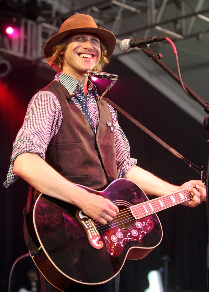 Todd Snider smiling while playing an acoustic guitar and harmonica on stage.