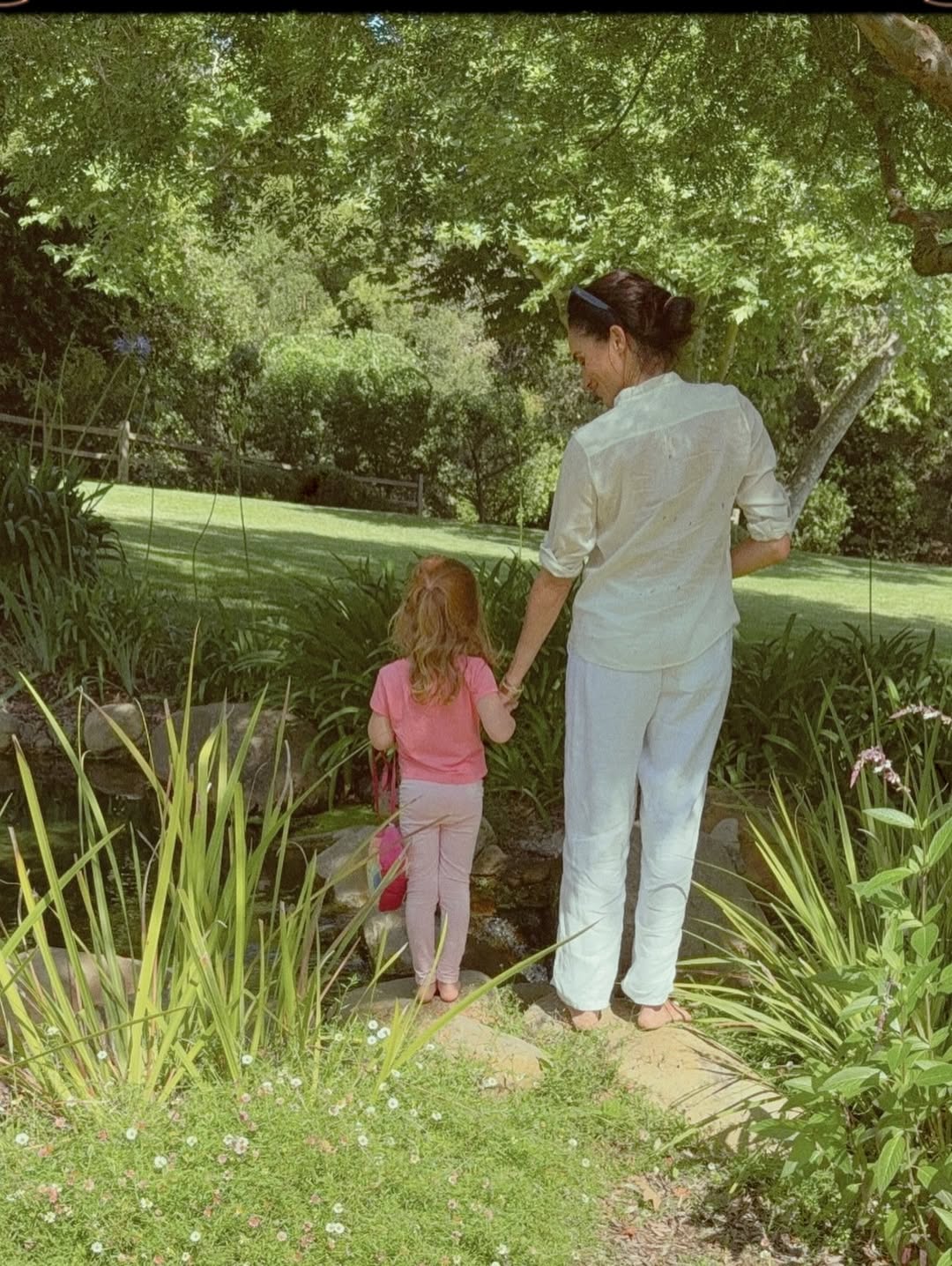 Meghan Markle holding a child's hand while standing by a small stream in a garden.