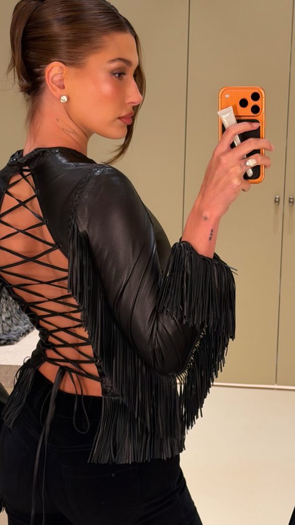 Hailey Bieber taking a mirror selfie in a black fringe and lace-up top.