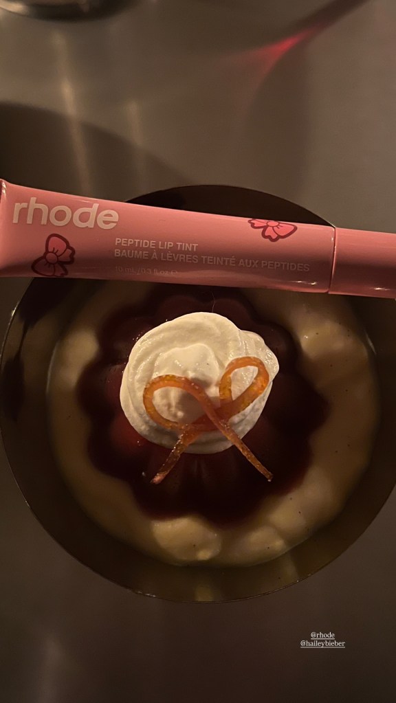 A pink Rhode Peptide Lip Tint tube next to a dessert with white cream, red jam, and candied orange peel.