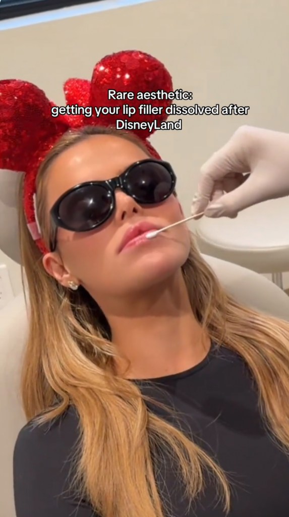 Brooks Nader at a medspa wearing red sequin Minnie Mouse ears while dissolving her lip filler.