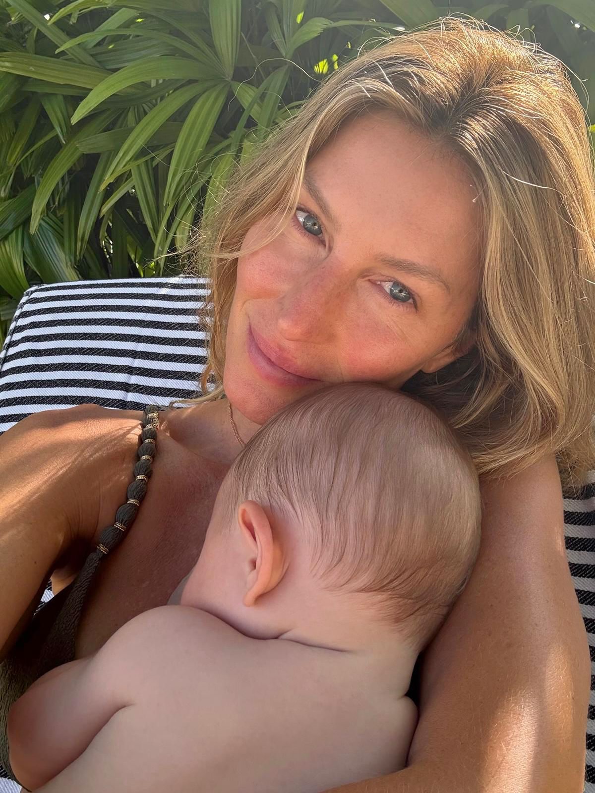 Gisele Bundchen smiles at the camera while holding a baby close to her chest.