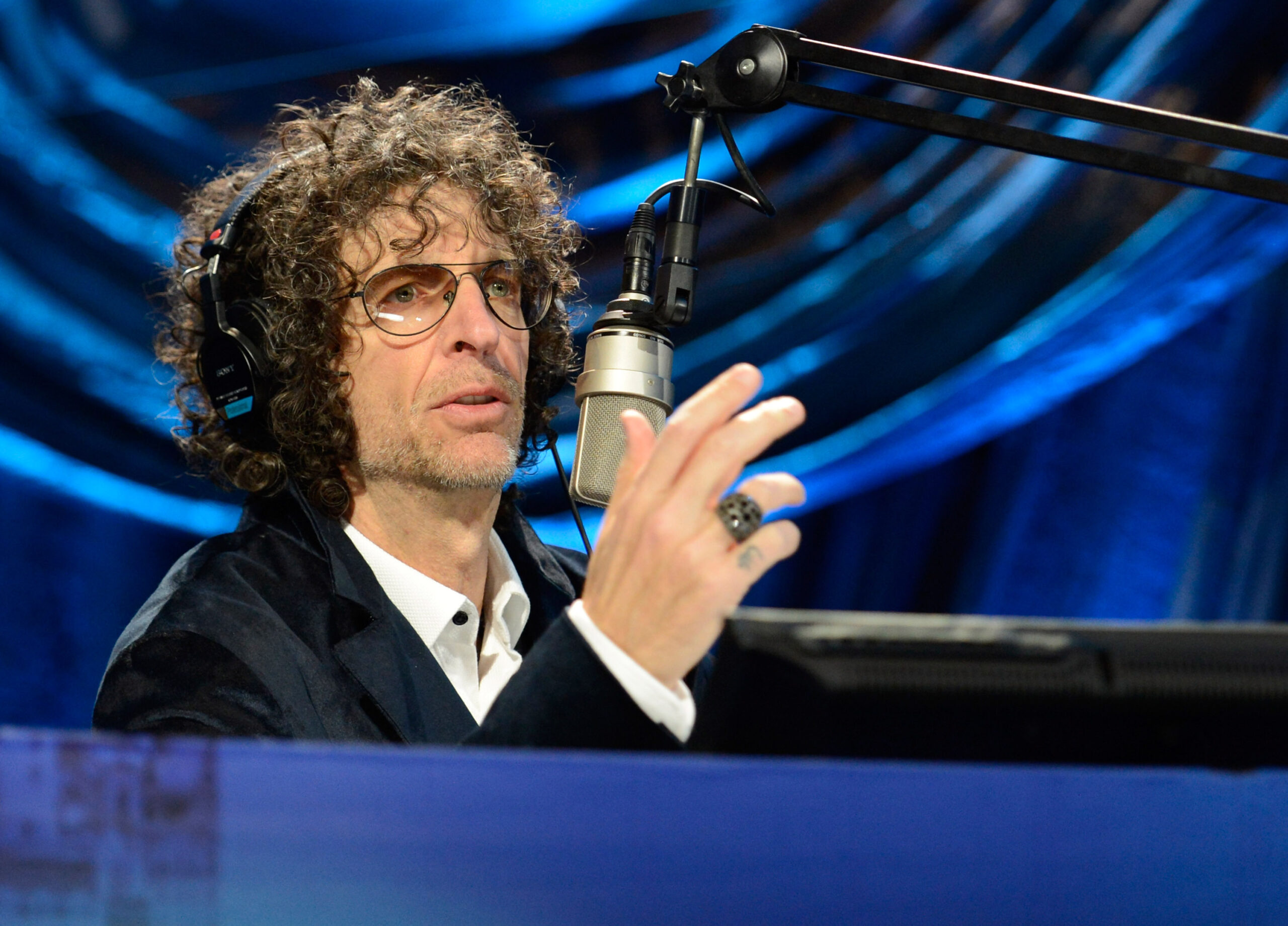 Howard Stern speaking on stage into a microphone after teasing a big announcement about his SiriusXM future.