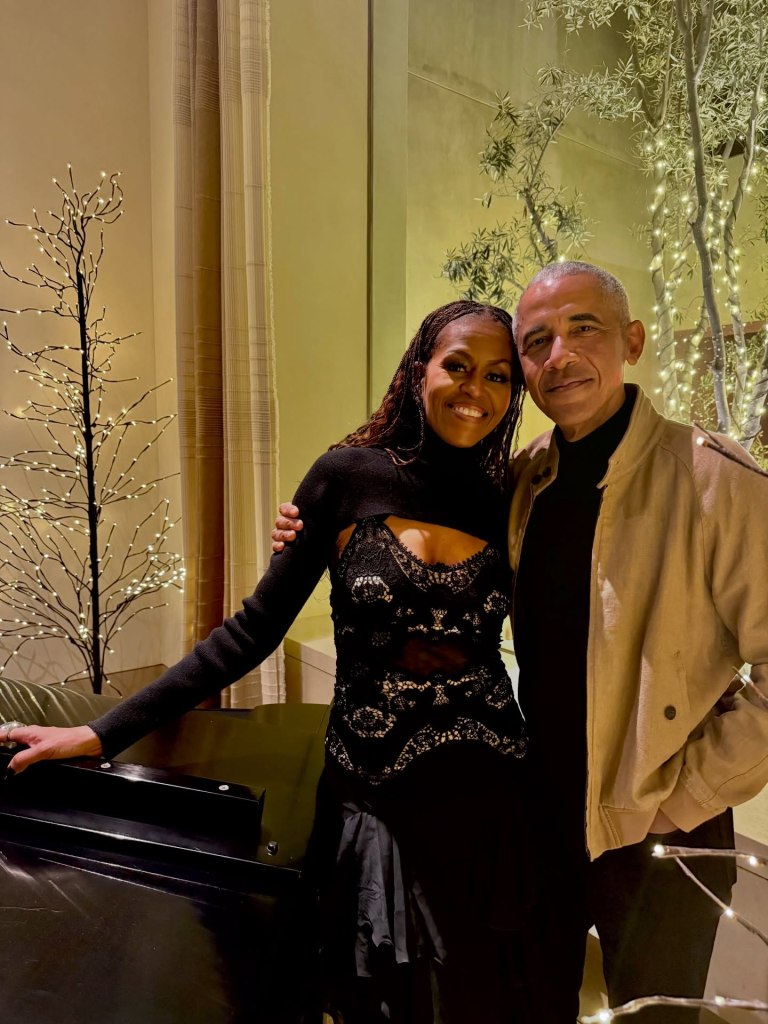 Michelle and Barack Obama stand together, smiling, with Christmas lights in the background.