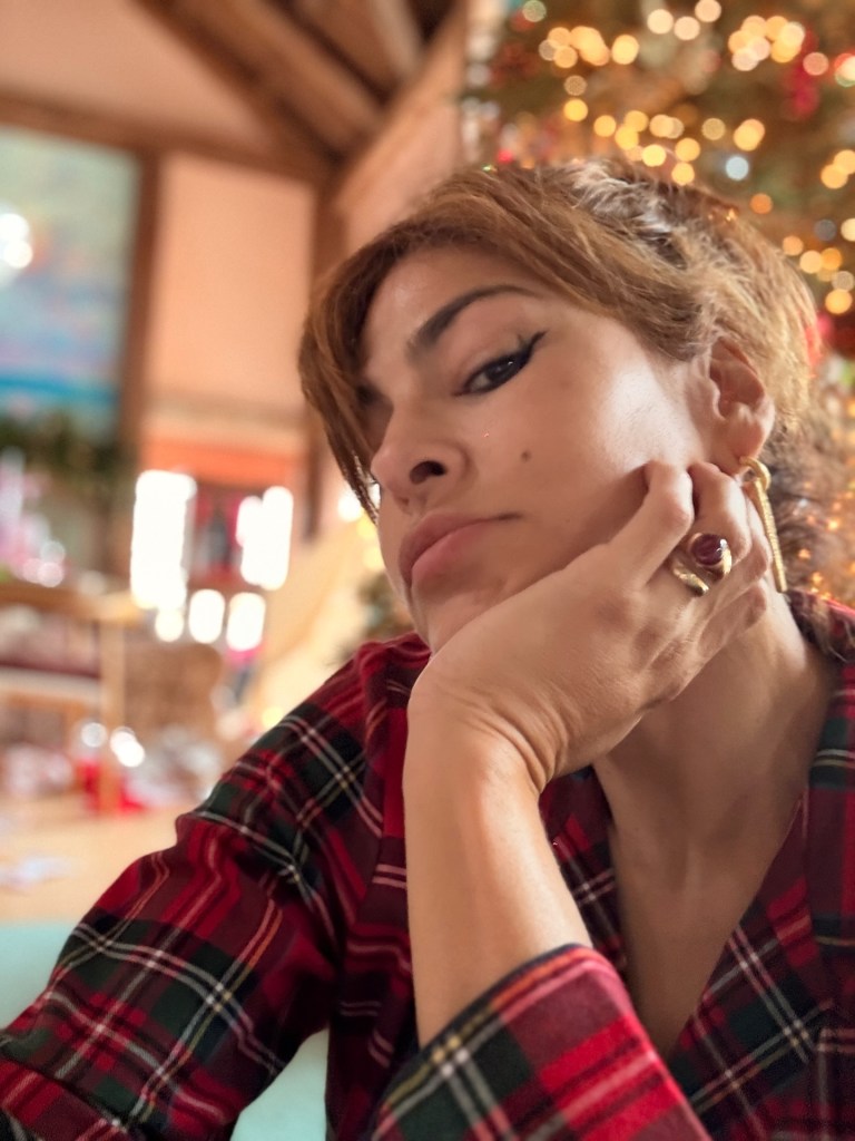 Eva Mendes wearing a red and green plaid shirt, resting her chin on her hand, with a Christmas tree in the background.