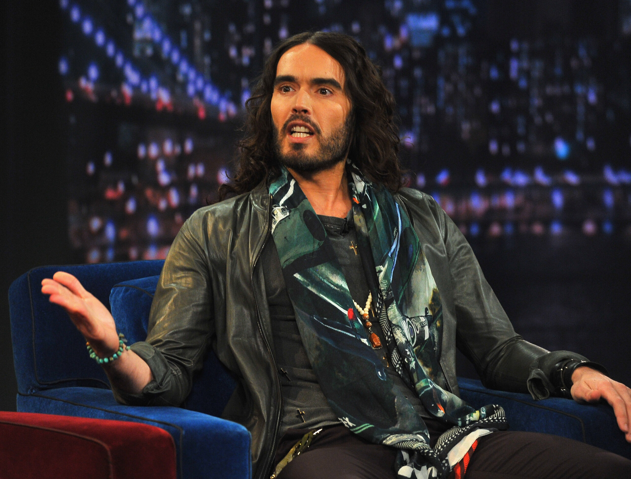 Russell Brand sitting on a blue couch, talking.