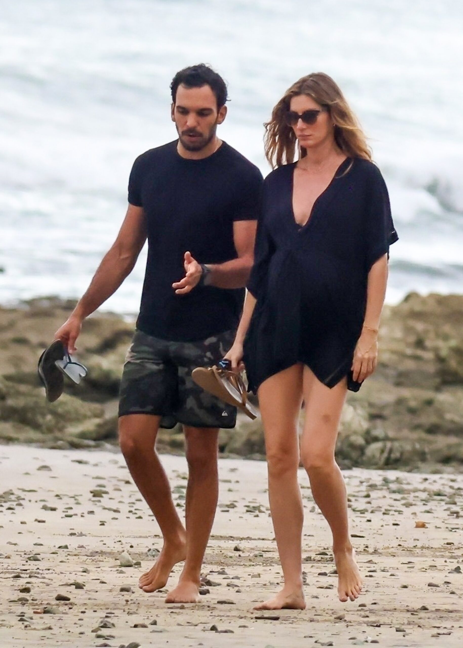 Gisele Bundchen and Joaquim Valente walking barefoot on a Miami beach, holding their sandals.