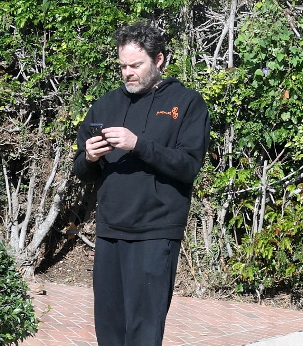 Bill Hader in a black hoodie and slide sandals, talking on his phone while walking in Los Angeles.