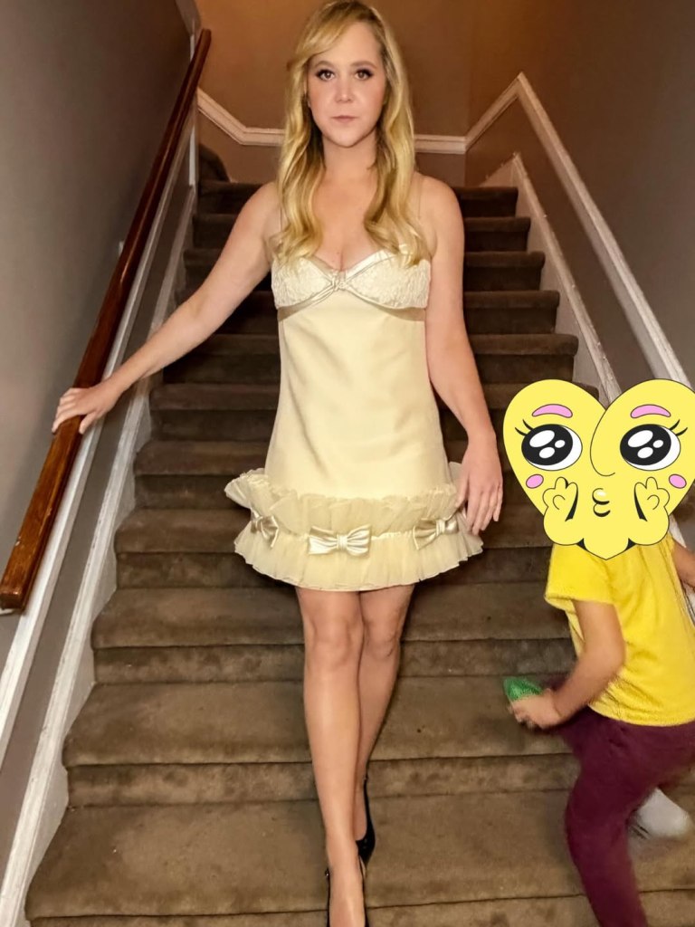 Amy Schumer poses on a staircase wearing a yellow lace minidress and black heels.