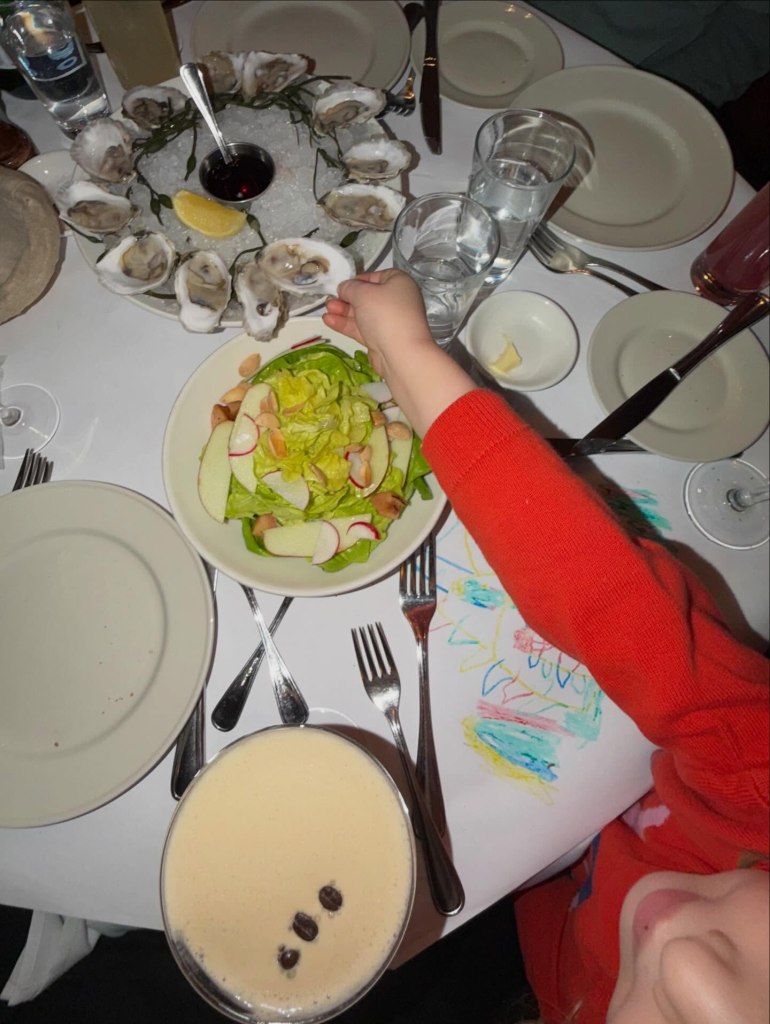 Overhead view of a holiday table with oysters, salad, drinks, and a child's arm in a red sweater reaching for an oyster.