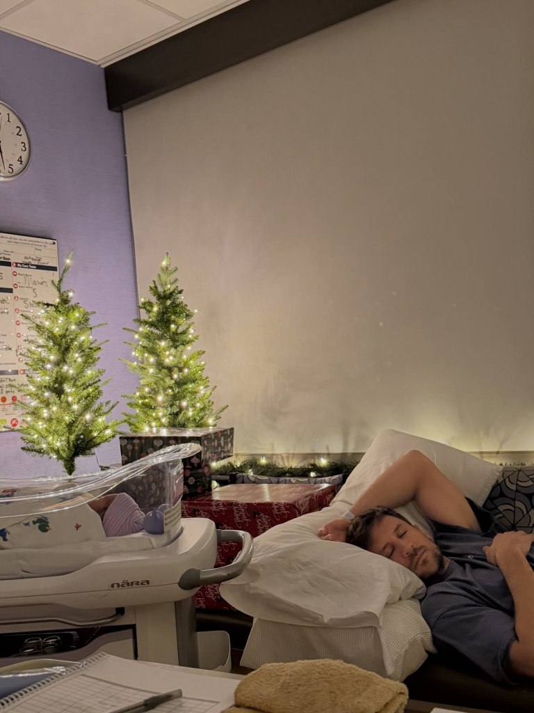 Newborn Romy Blair Rodgers sleeps in a hospital bassinet as Jordan Rodgers naps nearby, with small Christmas trees and wrapped gifts in the background.