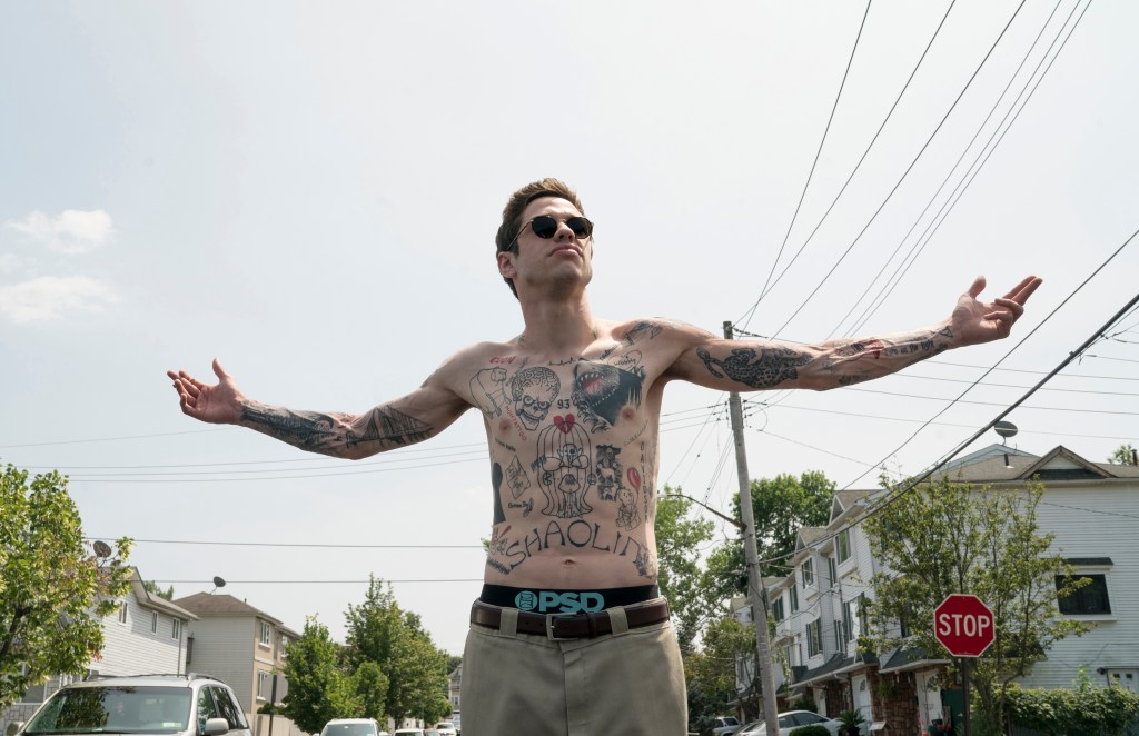 Pete Davidson stands shirtless outdoors with arms outstretched, showing the dramatic fading of his tattoos after removal treatments.