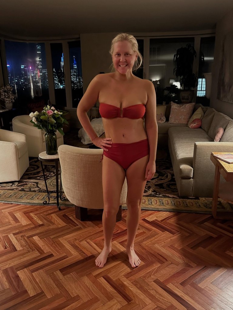 Amy Schumer posing in a red bikini in a living room with city views at night.