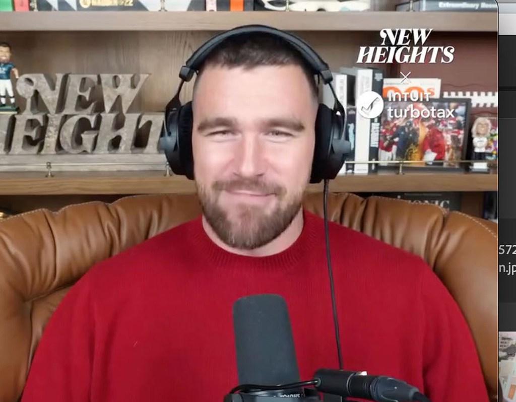 Travis Kelce on the New Heights podcast set, wearing a red sweater and headphones.