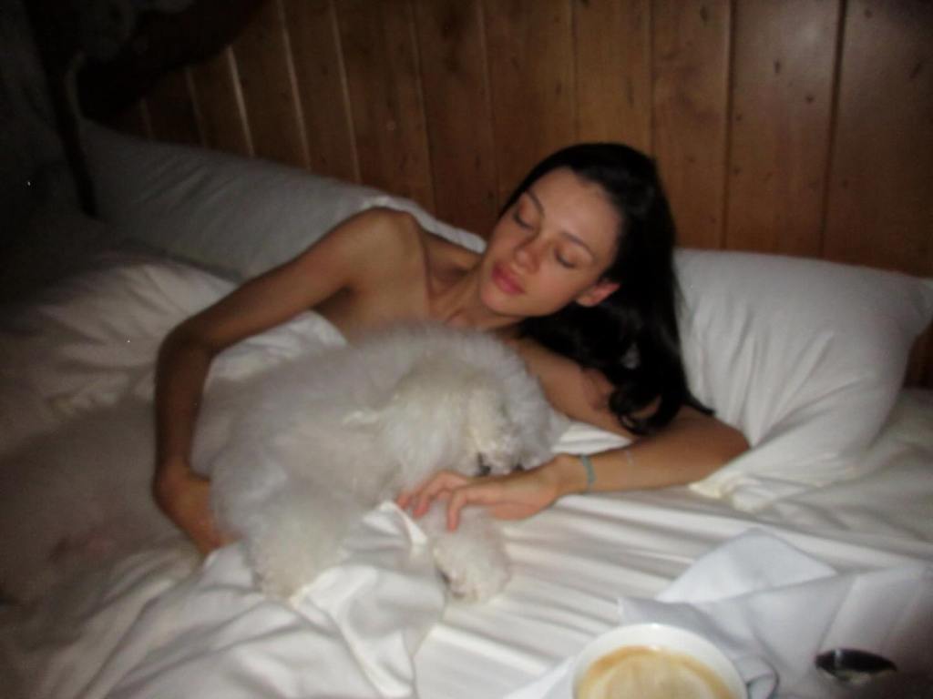 Nicola Peltz lying in bed under a white comforter, cuddling a dog; shared by Brooklyn Beckham on Instagram.