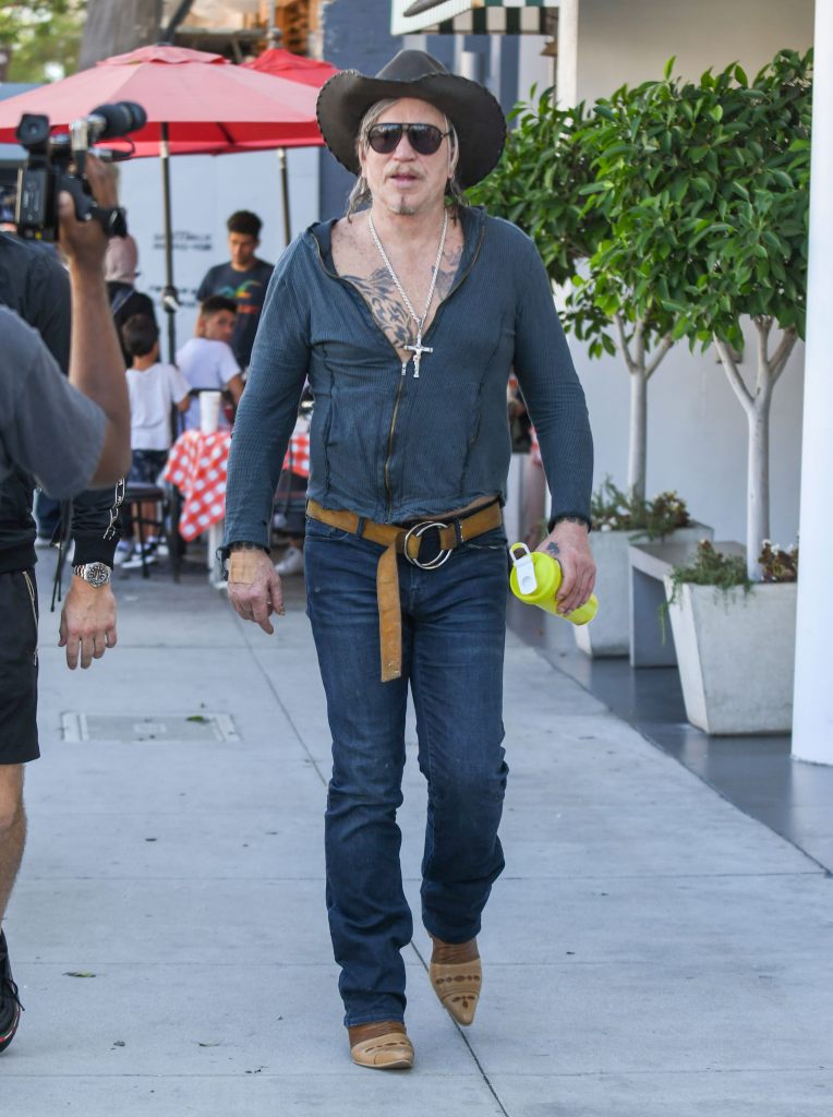 Mickey Rourke walking outdoors, wearing a black cowboy hat, sunglasses, a partially unzipped blue top, jeans, cowboy boots, and holding a yellow water bottle.