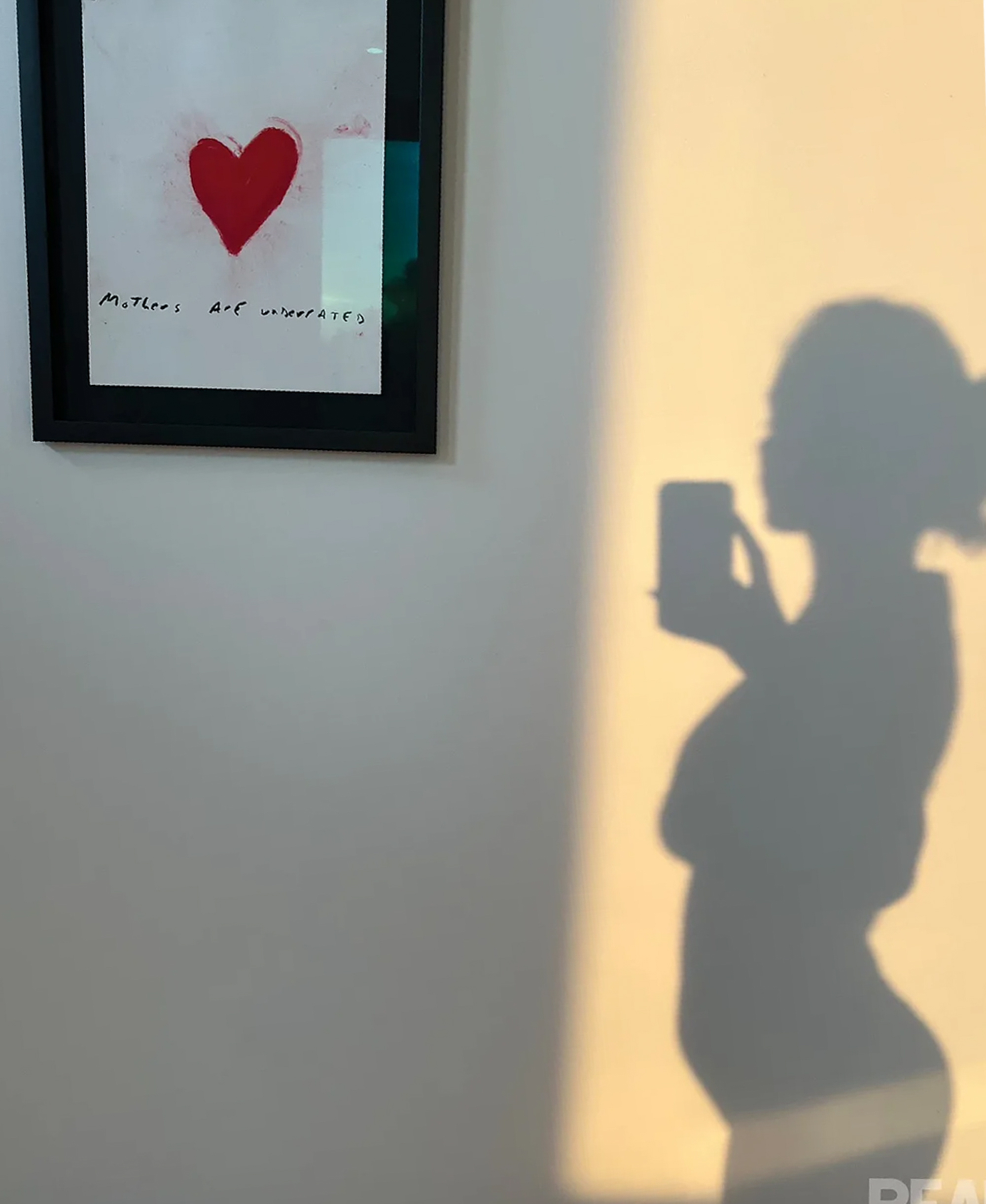 Shadow silhouette of Hailee Steinfeld showing a baby bump beside a framed red heart print that reads 'Mothers are underrated.'