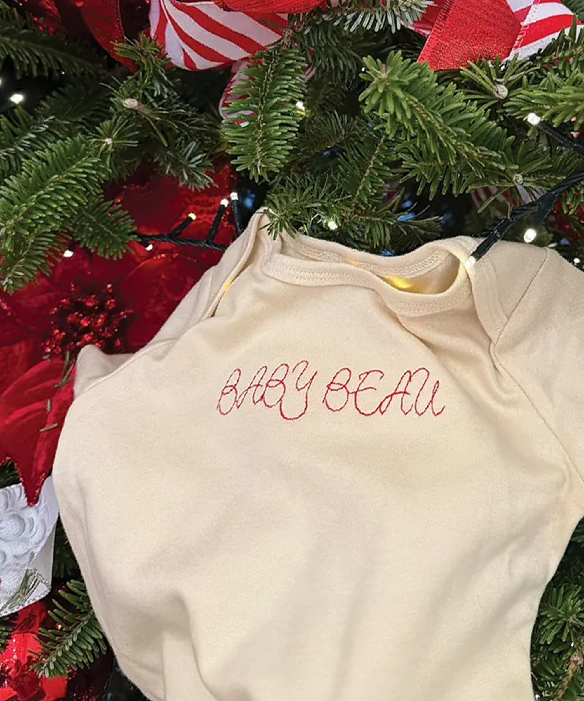 Cream baby onesie embroidered with 'BABY BEAU' in red, hanging on a Christmas tree.