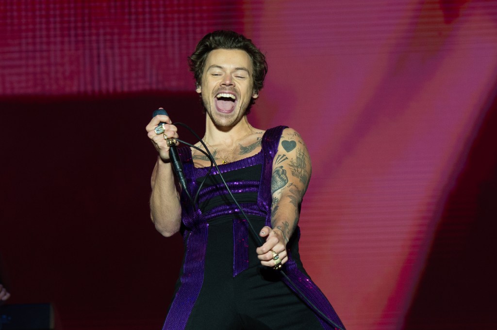 Harry Styles performs on stage with a microphone in a purple and black sequined outfit.
